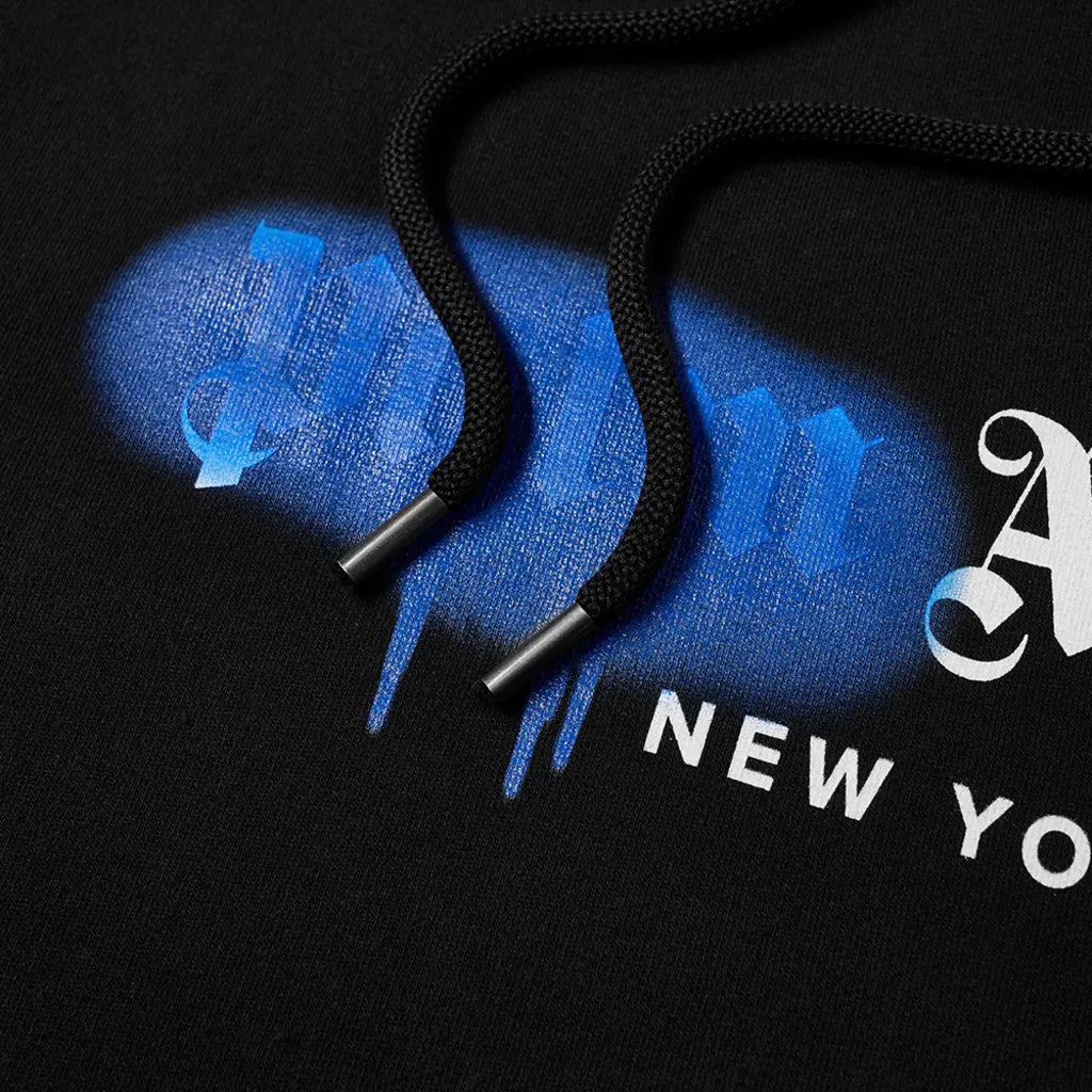Close-up of the Palm Angels New York Sprayed Logo Hoodie in black, highlighting its textured drawstrings. The fabric displays a distinctive New York sprayed logo with graffiti-style text in blue and white. The lighting accentuates the intricate details and texture of the design.