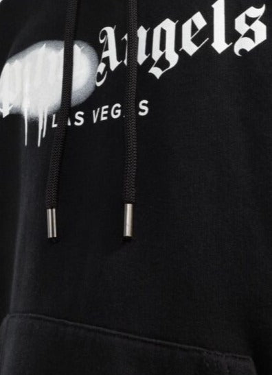 Close-up of the Palm Angels Las Vegas Sprayed Logo Hoodie showcasing Gothic-style text. The "Angels" word includes a drip effect over the "P", and "LAS VEGAS" appears below in smaller font. This striking Las Vegas Hoodie features black drawstrings with metal tips.