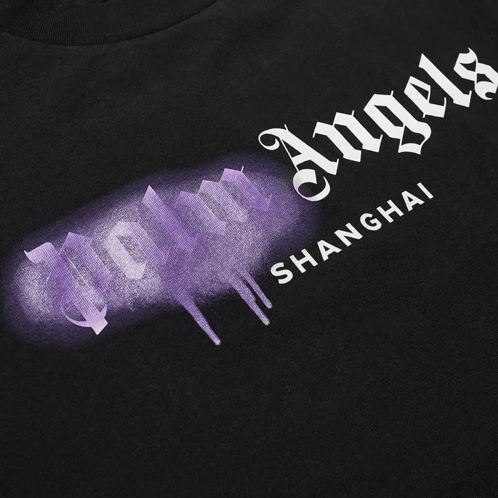 Close-up of the streetwear staple, the Palm Angels Shanghai Sprayed Logo T-Shirt, showcasing "Palm Angels" spray-painted in purple with a drip effect and "Shanghai" printed underneath in white.