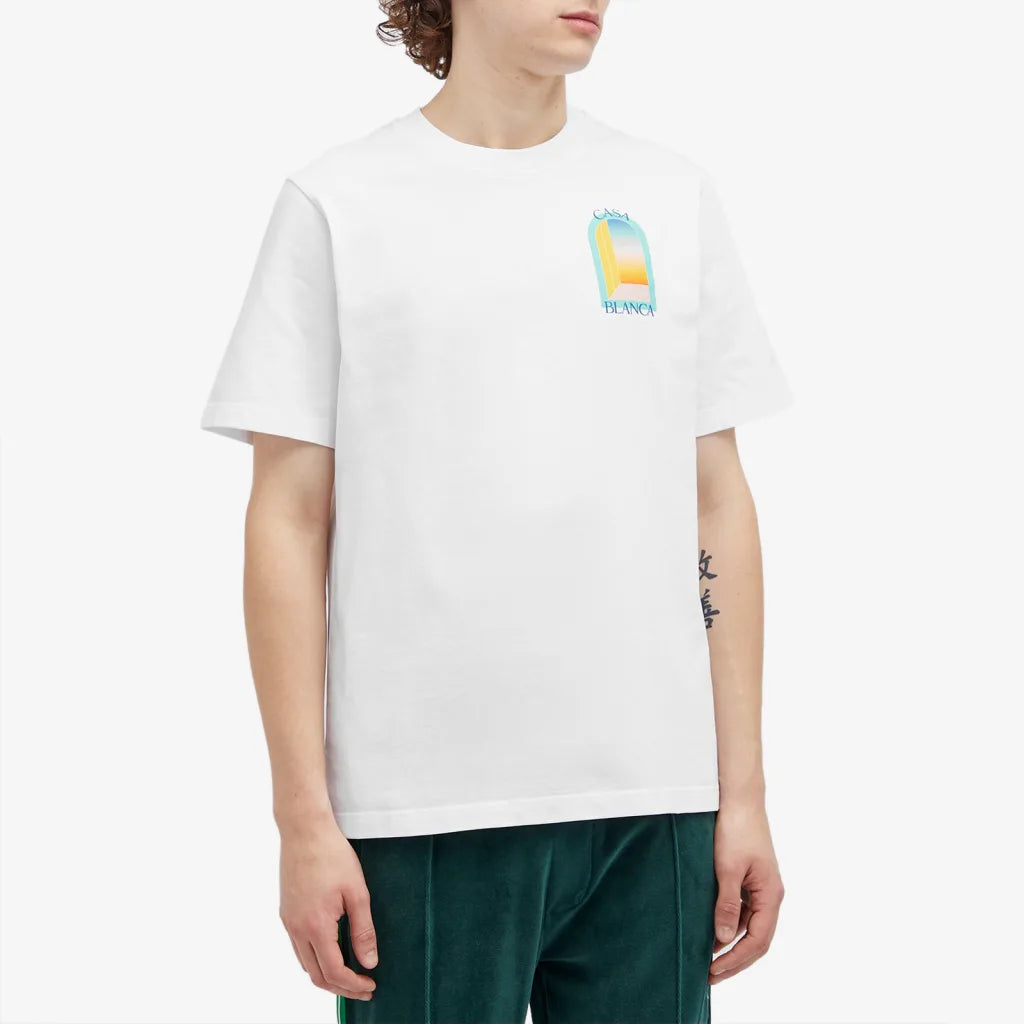 Someone in a vibrant Casablanca L'Arc Colore T-Shirt White, made from organic cotton, stands against a plain background. With curly hair and dark green pants, their forearm tattoo is visible.