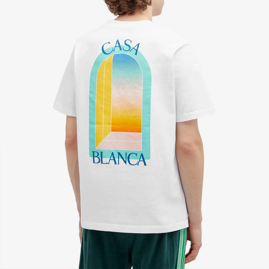 Someone is wearing a Casablanca L'Arc Colore T-Shirt in white, made from organic cotton, showcasing a colorful sunset arched window on the back with "CASA BLANCA" above and below the design. They pair it with dark green shorts for a complete look.