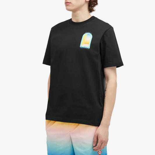 A person wears a black Casablanca L'Arc Colore T-Shirt, made from organic cotton with a vibrant chest graphic. They're paired with pastel gradient shorts shifting from pink to blue against a stark white backdrop that accentuates the colorful outfit.