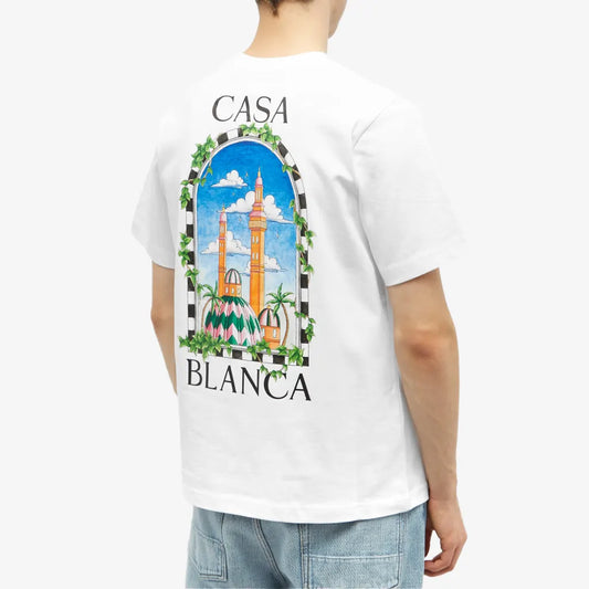 A person wearing the Casablanca Vue De Damas T-Shirt White, crafted from organic cotton, displays a mosque and palm trees in an arched window on the back. "CASA BLANCA" is elegantly printed around the design and paired with light blue denim jeans for a chic Afro-Cubism vibe.