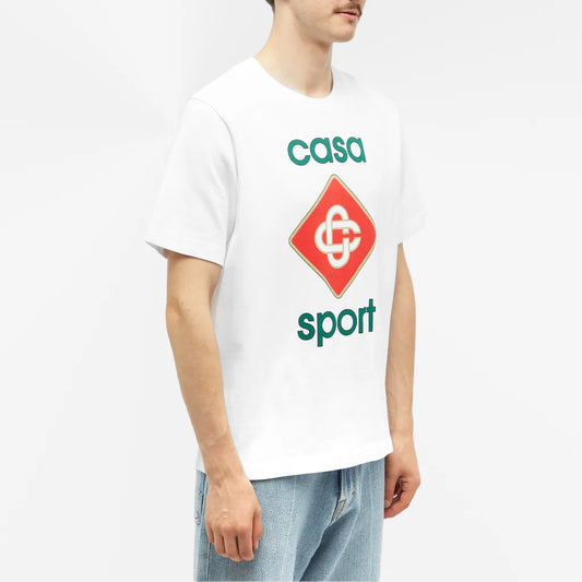 The person wears a Casablanca Casa Sport Logo T-Shirt in 100% organic cotton, with a red diamond logo and "casa sport" in green, paired with light blue jeans against a white backdrop, showcasing sustainable fashion.