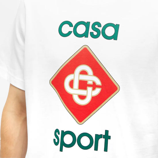 A person in a Casablanca Casa Sport Logo T-Shirt, made from 100% organic cotton, wears a white shirt with a red diamond-shaped logo featuring interlocking white letters and "casa sport" printed in green.