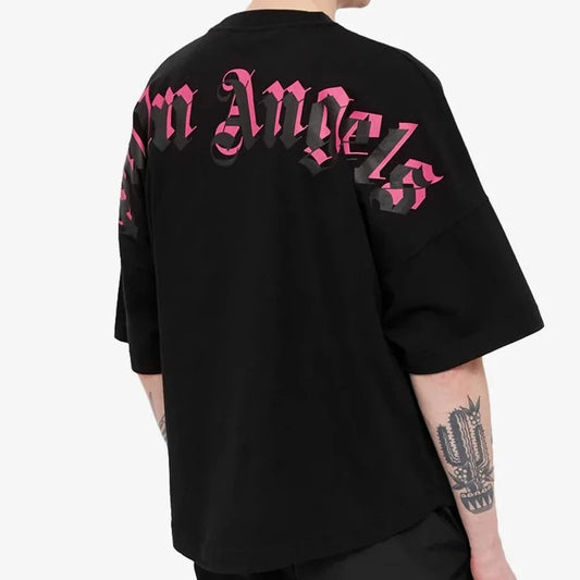 The individual is dressed in a Palm Angels Oversize Double Printed Neck Logo T-Shirt in black and pink, featuring Gothic-style text and a logo on the back. This soft cotton shirt enhances their short hair and tattooed left forearm against a plain white background.