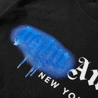 The Palm Angels New York Sprayed Logo T-Shirt features a close-up of a black fabric with a blue spray paint design and stylized text. Below, white text spells out "New York." The blue design showcases a gradient effect with drips at the bottom, capturing streetwear vibes reminiscent of Palm Angels' bold aesthetic.