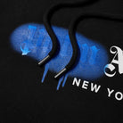 Close-up of the Palm Angels New York Sprayed Logo Hoodie in black, highlighting its textured drawstrings. The fabric displays a distinctive New York sprayed logo with graffiti-style text in blue and white. The lighting accentuates the intricate details and texture of the design.