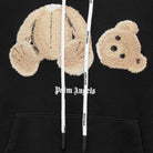 The Palm Angels Bear Hoodie features a fluffy teddy bear graphic with its head and body separated, embodying high fashion streetwear. The phrase "Palm Angels" is prominently printed below, while the drawstrings include additional text details.