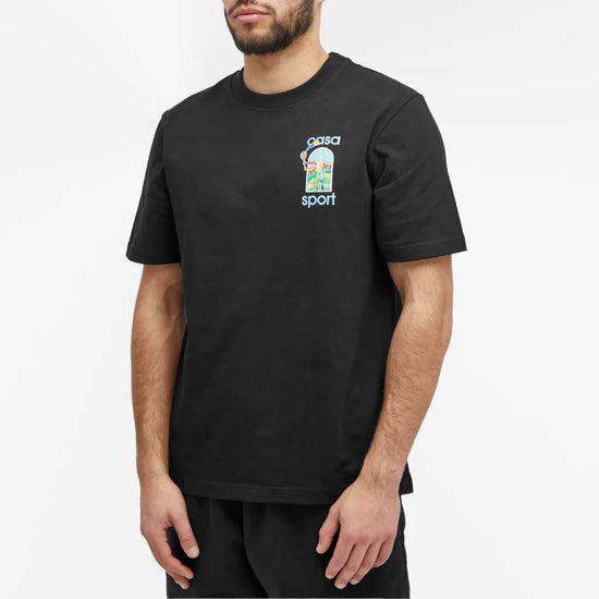 A person wears a Casablanca Le Jeu Colore T-Shirt Black featuring a colorful graphic and "casa sport" text, standing against a plain white background. Made from organic cotton, the shirt pairs perfectly with black pants for a sleek look.