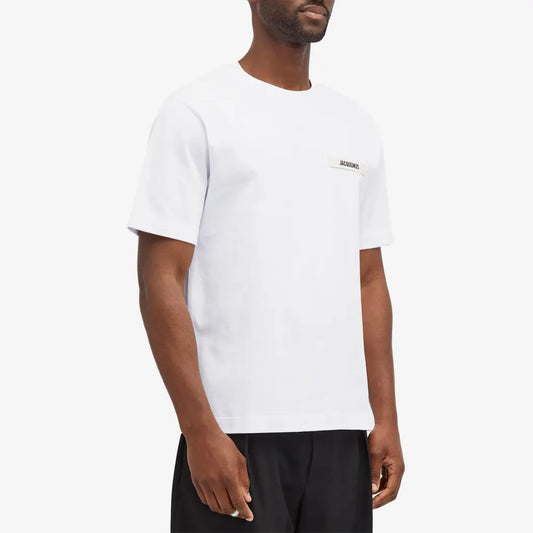 A person models a Jacquemus Gros Grain Logo T-Shirt in white with organic cotton and embroidered logo against a plain background, paired with dark pants, face not shown.