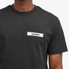 Someone is wearing a Jacquemus Gros Grain Logo T-Shirt Black made from organic cotton fleece. It has a small white patch with an embroidered grosgrain logo on the front, set against a plain white background.