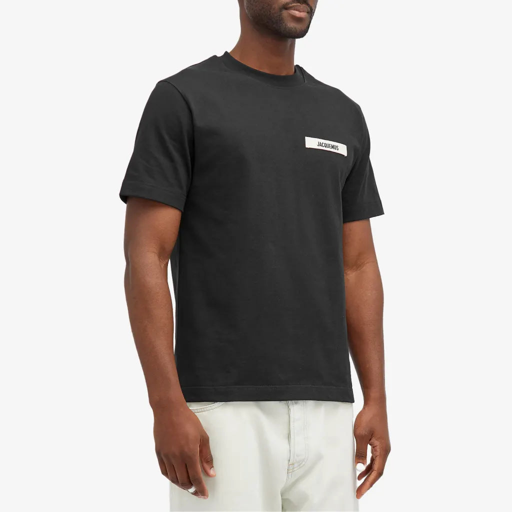 A man wearing a Jacquemus Gros Grain Logo T-Shirt Black stands against a plain background, the embroidered grosgrain logo on his chest enhancing his style. The black shirt contrasts with his light-colored pants for a modern and stylish look.