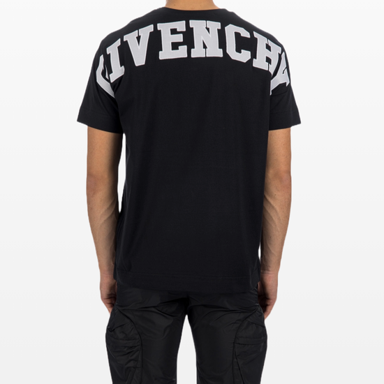 A person sporting a Givenchy Paris Eiffel College Logo T-Shirt with bold branding on the back stands facing away, showcasing their casual wardrobe. They also wear black cargo pants. The plain white background emphasizes the chic simplicity of the ensemble from Givenchy.