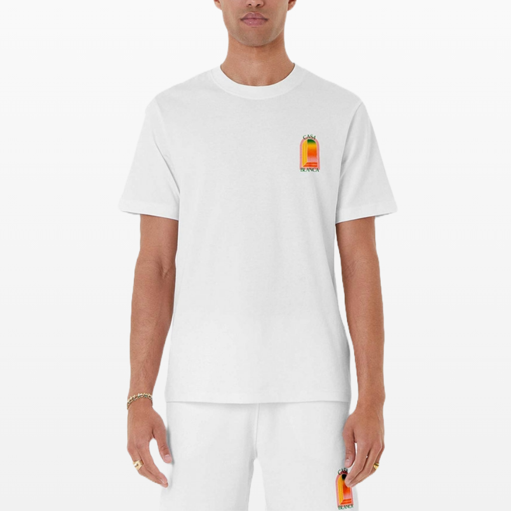 A person wearing a Casablanca Gradient Arch Logo T-Shirt White in organic cotton stands against a plain background. The t-shirt features vibrant hues and a small, colorful patch on the left side, adding a playful touch to the ensemble.