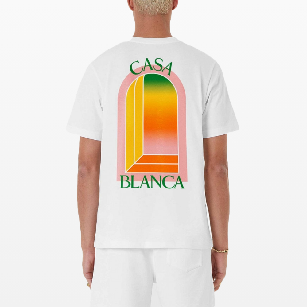 A person is seen from the rear wearing a Casablanca Gradient Arch Logo T-Shirt White made of organic cotton, paired with shorts. The shirt features vibrant colors and a geometric arch adorned with "CASA BLANCA" in green text.