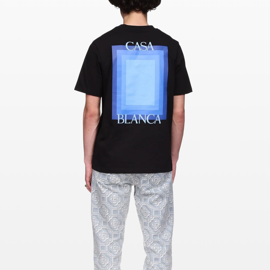 Someone is wearing a black Casablanca Logo Degrade Screen Printed T-Shirt featuring a blue geometric design and "CASABLANCA" text on the back, complemented by light-colored patterned pants. The outfit contrasts with a plain white background.