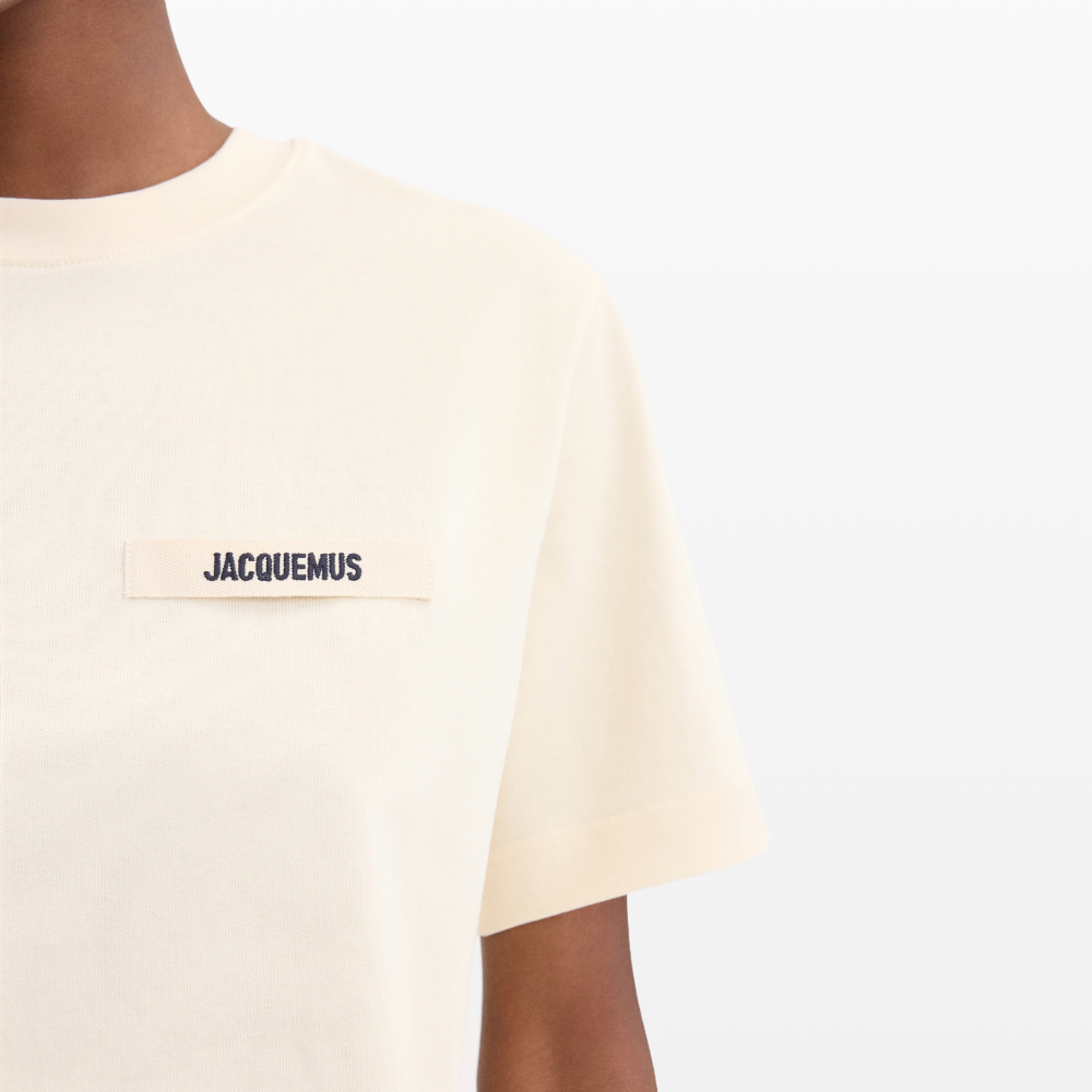 Close-up of a person in a Jacquemus Gros Grain Logo T-Shirt Beige, made of organic cotton. The shirt displays an embroidered grosgrain logo on the chest, with the image emphasizing the label and upper part against a softly blending background.