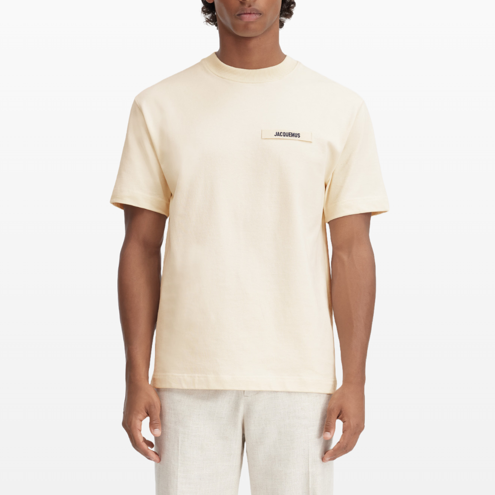 A person stands against a plain white background wearing a Jacquemus Gros Grain Logo T-Shirt Beige made of organic cotton, featuring an embroidered grosgrain logo on the chest, paired with cream-colored pants.