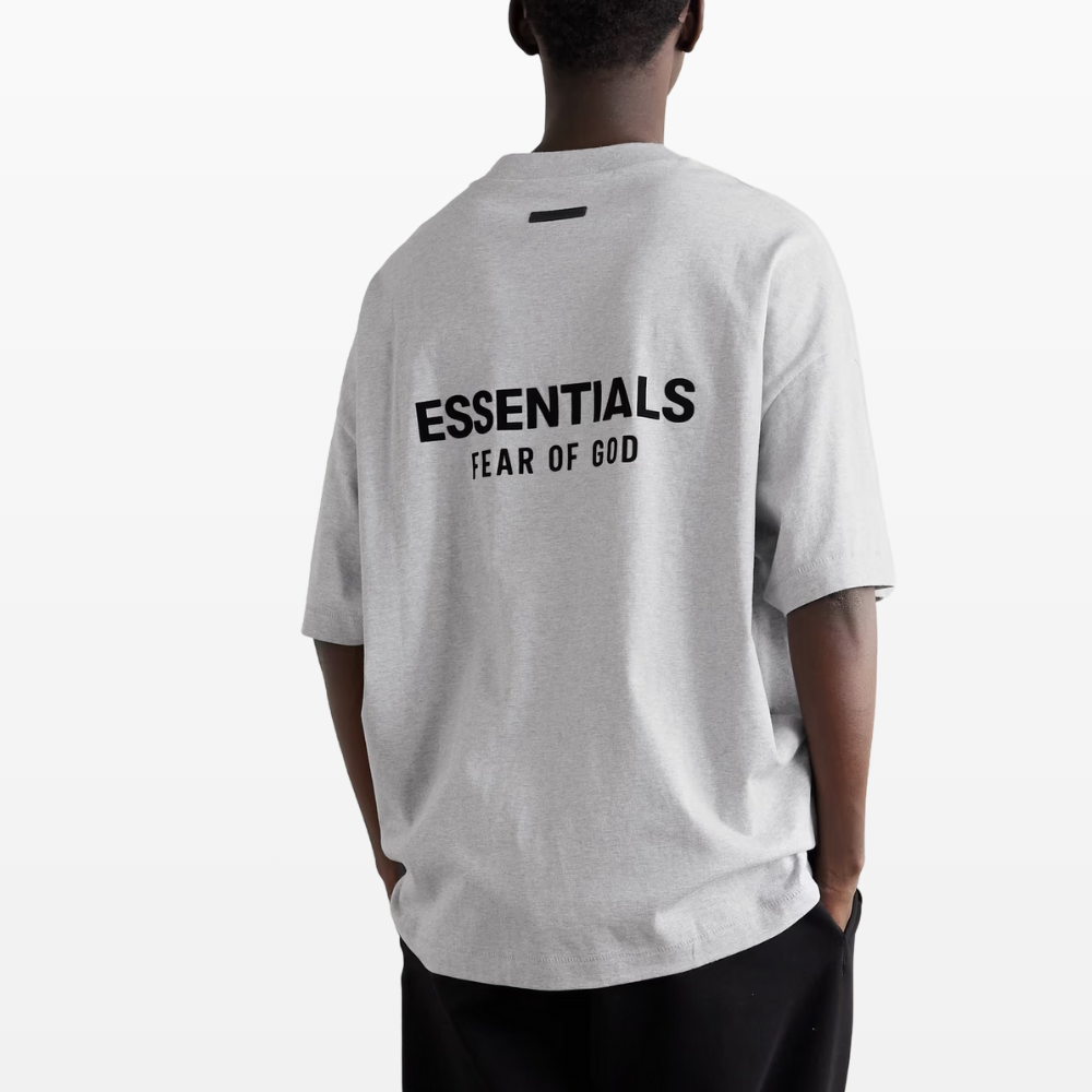 The individual is wearing a light heather grey Fear of God ESSENTIALS HOLIDAY24 T-Shirt, crafted from 100% cotton jersey, featuring "FEAR OF GOD ESSENTIALS" printed in bold black on the back. They're standing against a plain white background, facing away and paired with dark pants.