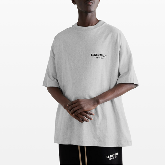 A person wears the Fear of God ESSENTIALS HOLIDAY24 T-Shirt in Light Heather Grey, crafted from 100% cotton jersey with the "ESSENTIALS Fear of God" logo, paired with black shorts featuring the same design, all set against a plain white background.