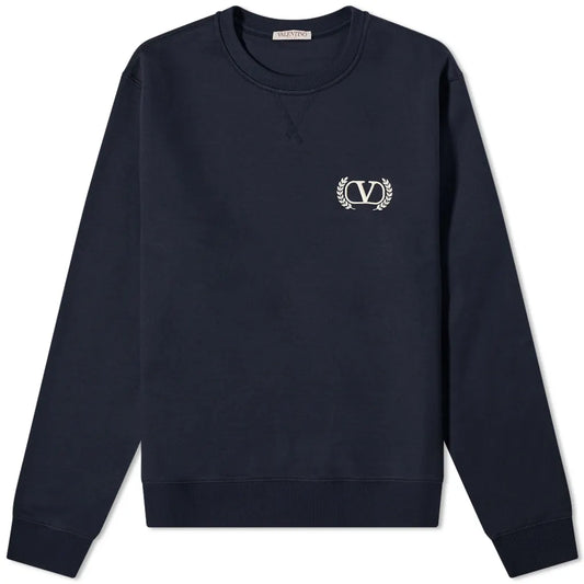 The Valentino Varsity V Logo Sweatshirt is a luxurious garment featuring a deep blue color and an embroidered white emblem on the left chest. Its long sleeves are adorned with circular designs and laurel leaves, while ribbed cuffs and hem complete its sophisticated appearance.