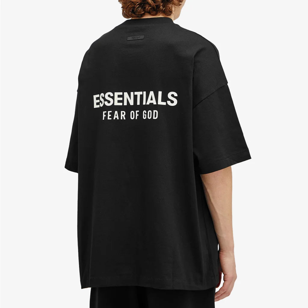 A person in a black Fear of God ESSENTIALS HOLIDAY24 T-Shirt with "ESSENTIALS FEAR OF GOD" in white on the back, embodying fashion-forward style against a plain white background.