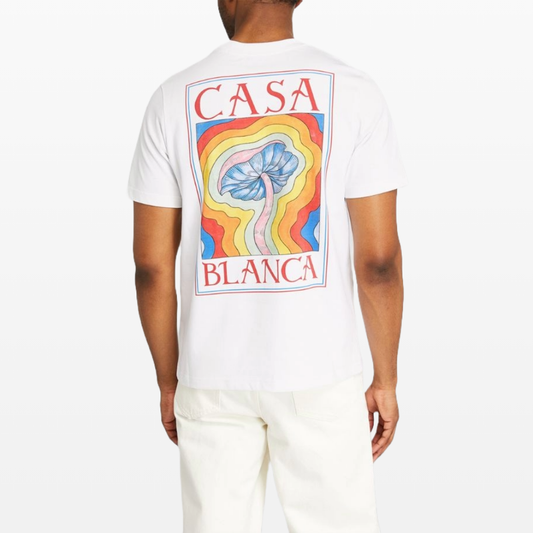 A person is sporting a Casablanca Paris Mind Vibrations T-Shirt crafted from organic cotton. It features vibrant abstract art on the back, showcasing mushroom graphics, a blue flower, and wavy rainbow-colored lines. The shirt is emblazoned with "Casa Blanca" and pairs seamlessly with their white pants.