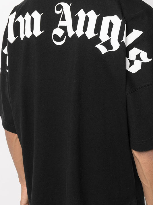 A person is shown from behind wearing the Palm Angels Oversize Neck Logo T-Shirt in black, featuring bold white Gothic lettering that partially reveals "lm Angels." This shirt showcases a striking logo print across the upper back and is crafted from soft cotton for a blend of style and comfort against a plain background.