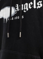 Close-up of the Palm Angels Las Vegas Sprayed Logo Hoodie showcasing Gothic-style text. The "Angels" word includes a drip effect over the "P", and "LAS VEGAS" appears below in smaller font. This striking Las Vegas Hoodie features black drawstrings with metal tips.