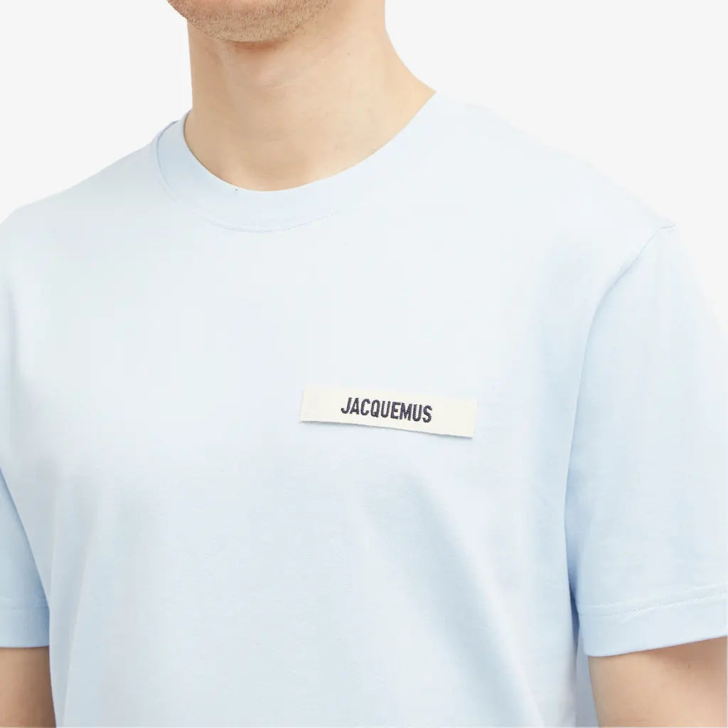 A person is wearing the Jacquemus Gros Grain T-Shirt in light blue with a rectangular "JACQUEMUS" patch on the chest against a plain white background.