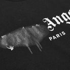 Close-up of a black Palm Angels Paris Sprayed Logo T-Shirt featuring white graffiti-style text. The iconic design is accentuated by the Palm Angels branding, with a portion of the text appearing smeared or faded to enhance the artistic streetwear style against the dark fabric.