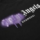 Close-up of the streetwear staple, the Palm Angels Shanghai Sprayed Logo T-Shirt, showcasing "Palm Angels" spray-painted in purple with a drip effect and "Shanghai" printed underneath in white.
