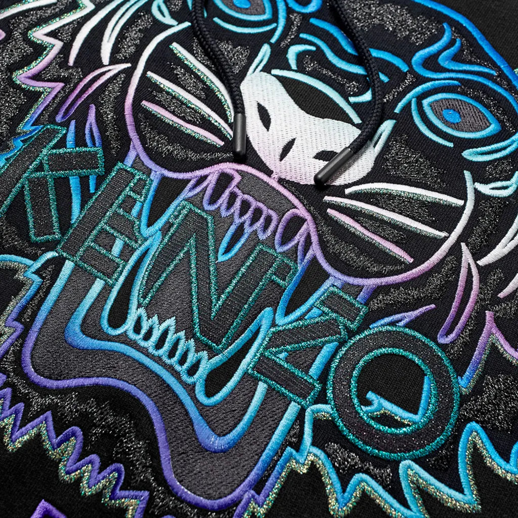 Close-up of a colorful embroidered tiger design on fabric. The intricate pattern features bold blue, purple, and white outlines with the word "KENZO" integrated into the tiger's open mouth, set against a black background on the Kenzo Holiday Capsule Limited Edition Tiger Embroidered Hoodie.