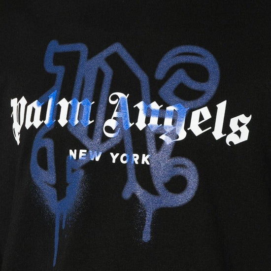 A black t-shirt from Palm Angels, the "New York Monogram Sprayed Logo T-Shirt," features the iconic "Palm Angels" name in a white Gothic-style font with "New York" elegantly inscribed underneath. Abstract blue patterns add a graffiti effect for a modern twist on the design.