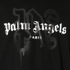 The Palm Angels Paris Monogram Sprayed Logo T-Shirt showcases a black background adorned with the brand's name "Palm Angels" elegantly scripted in white Gothic lettering, with "Paris" subtly placed underneath. Shadowy, dark patterns reminiscent of a monogram logo tee emerge behind the text, all crafted from high-quality premium cotton jersey.