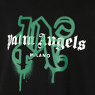 The image showcases a black fabric from the Palm Angels brand, specifically the Milano Monogram Sprayed Logo T-Shirt, which prominently features the monogram in a white, Gothic-style font set against a striking green spray-painted design.