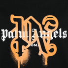 A close-up of the Palm Angels Roma Monogram Sprayed Logo T-Shirt features stylized white "Palm Angels" text over an orange, graffiti-style letter "P" with paint drips. Below it reads "Roma" in smaller white letters, all set against a black background.