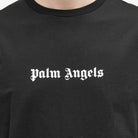 Close-up of a person wearing the Palm Angels Logo T-Shirt in black, featuring the brand name "Palm Angels" in white Gothic font at the center. The minimalist design effortlessly captures the essence of LA skater style.