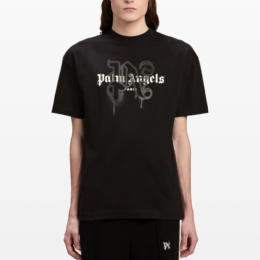 The individual is wearing a Palm Angels Paris Monogram Sprayed Logo T-Shirt, featuring the brand's name and city in a graffiti-style font on black cotton jersey. They pair it with matching black pants against a plain white background.