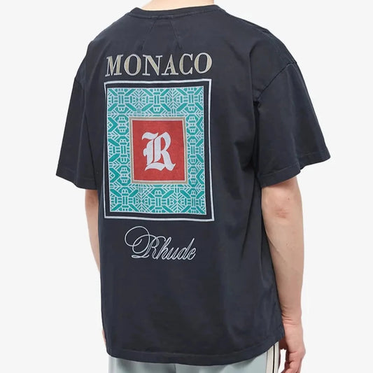 A person wearing the Rhude Monaco T-Shirt features signature streetwear design elements, including white "MONACO" lettering above a striking red square with a centered white "R," surrounded by teal patterns. Below the square, "Rhude" is tastefully scripted. The individual embodies a racing spirit and faces away.