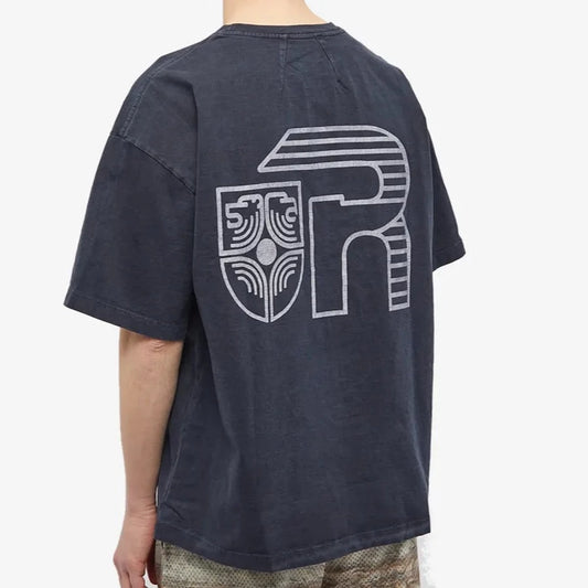 A person is dressed in a vintage black Rhude Banque De Rhude T-Shirt by Rhude, showcasing a large patterned design on the back with a stylized letter "R" and an ornate emblem. This 100% cotton tee is teamed with camouflage-patterned shorts.