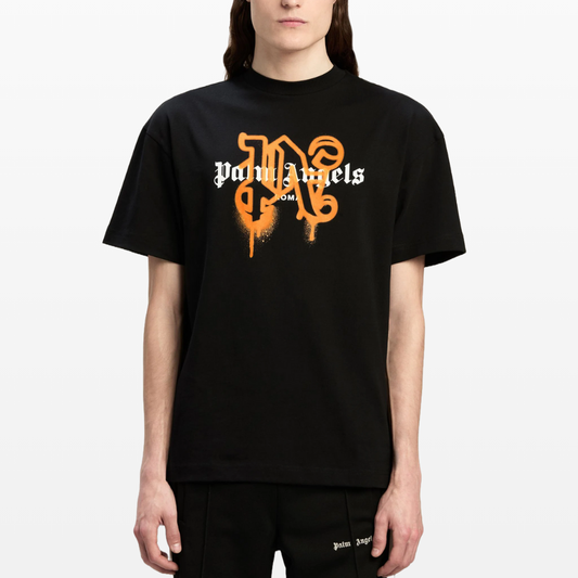 A person wearing a Palm Angels Roma Monogram Sprayed Logo T-Shirt, featuring orange and white stylized lettering on the front. This black tee, crafted from soft cotton jersey, has short sleeves and is paired with black pants. The background is plain white.