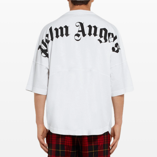 Rear view of a person wearing the Palm Angels Oversize Neck Logo T-Shirt in white, showcasing the brand's signature "Palm Angels" logo printed in black gothic font on the back. They pair it effortlessly with red and black plaid pants, embracing the relaxed oversized fit.