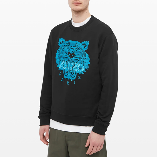 A person is showcasing an urban style with the Kenzo Neon Tiger Embroidered Sweatshirt, featuring a prominent blue embroidered "KENZO" and "PARIS" design. Completing the look are an underlying white shirt and olive green pants.