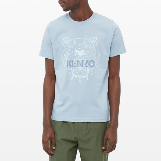 A man dons the Kenzo Classic Festive Tiger T-Shirt, showcasing the brand's iconic tiger logo on a light blue luxe cotton tee. He pairs it stylishly with green pants.