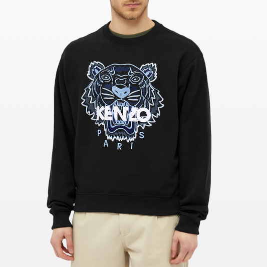 A man is showcasing the Kenzo Classic Tiger Embroidered Sweatshirt, distinguished by its large, embroidered tiger emblem and "KENZO PARIS" text on the front. He complements it with beige pants, adding a hint of Parisian elegance. The scene is set against a plain white background.