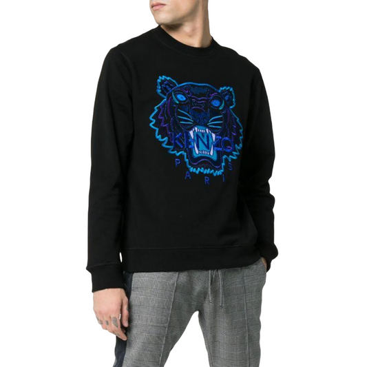 A person wears the Kenzo Holiday Capsule Embroidered Sweatshirt in black, adorned with a striking tiger design that makes it ideal for a winter wardrobe, complemented by gray plaid pants. The backdrop is white.