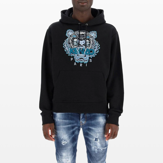 A person is wearing a Kenzo Classic Tiger Embroidered Hoodie in black, showcasing the "Paris" branding and featuring a kangaroo pouch pocket. They have paired it with distressed blue jeans for a stylish yet casual look.