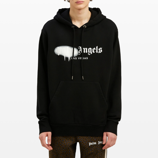 A person wearing the Palm Angels Las Vegas Sprayed Logo Hoodie in black, adorned with white "Angels Las Vegas" text and a paint drip design. The hoodie includes a front pocket. They are also sporting brown leopard print pants against a plain white background.